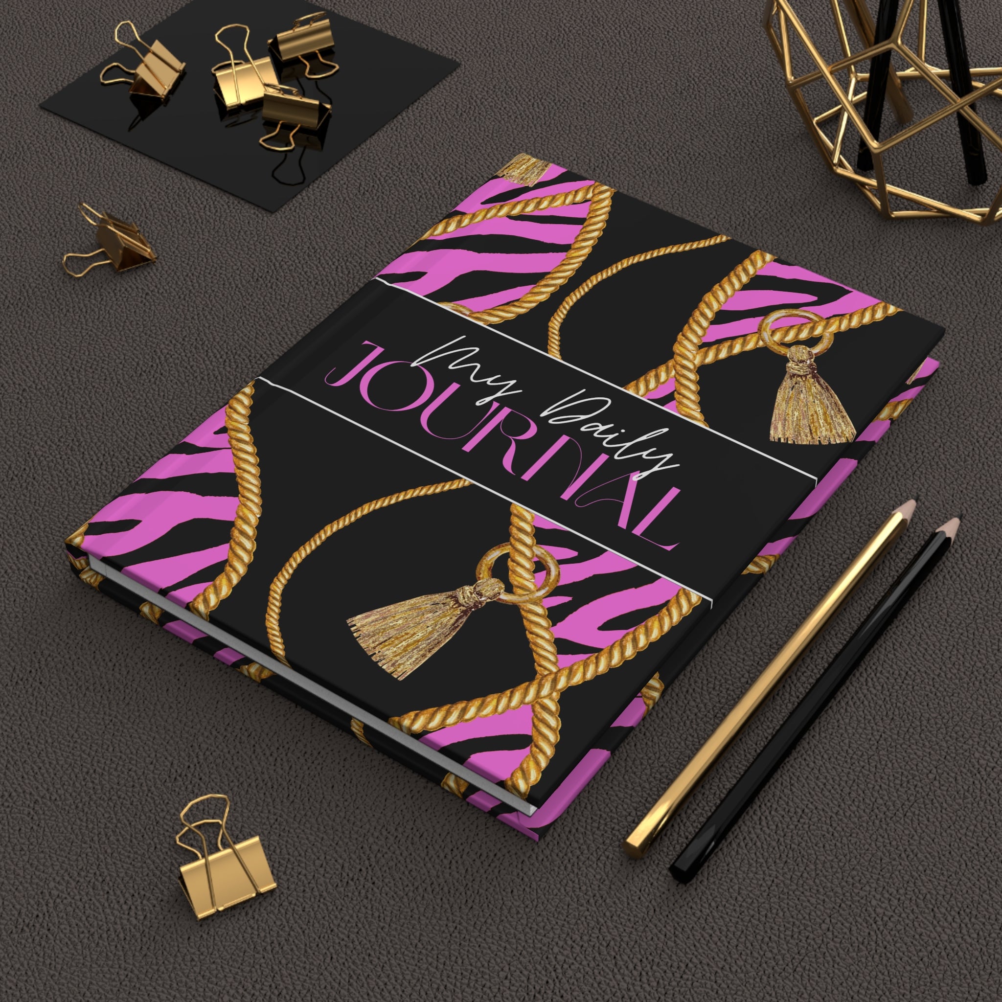 A hardcover journal with a black and pink zebra print cover decorated with gold rope and tassel motifs, styled on a dark textured surface with gold and black office accessories.
