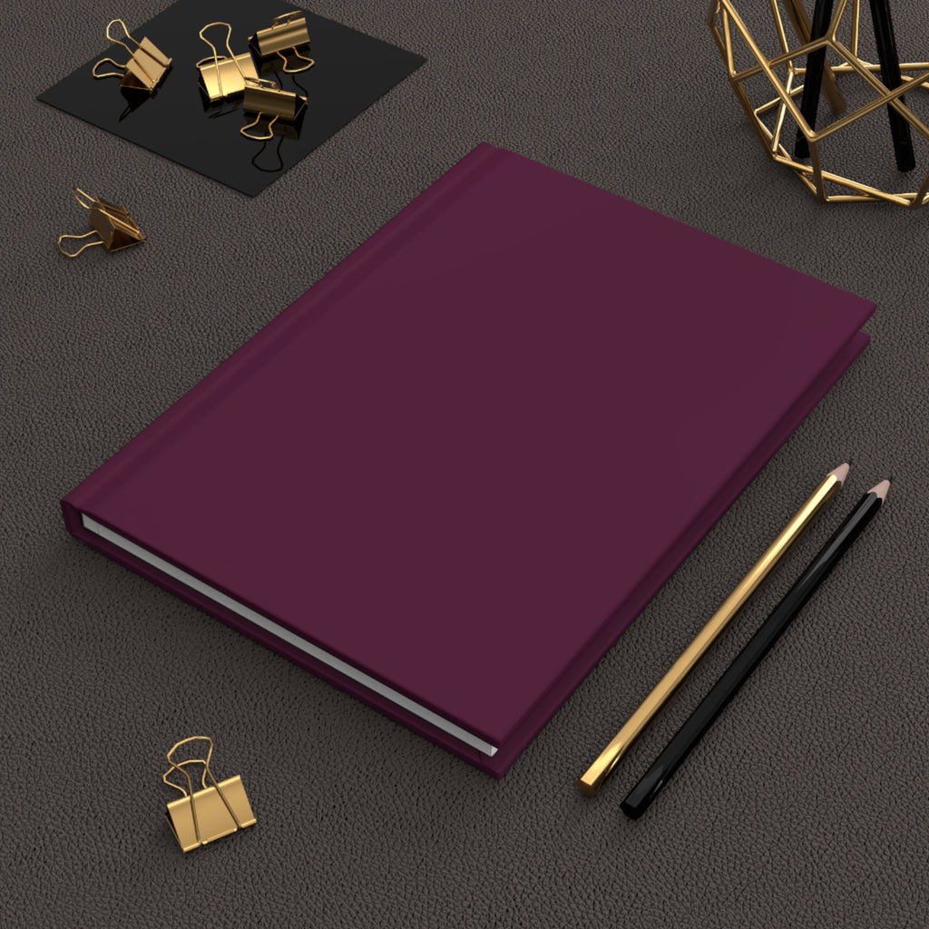 A hardcover notebook or journal in deep plum purple, styled on a textured dark gray surface with gold and black office accessories.