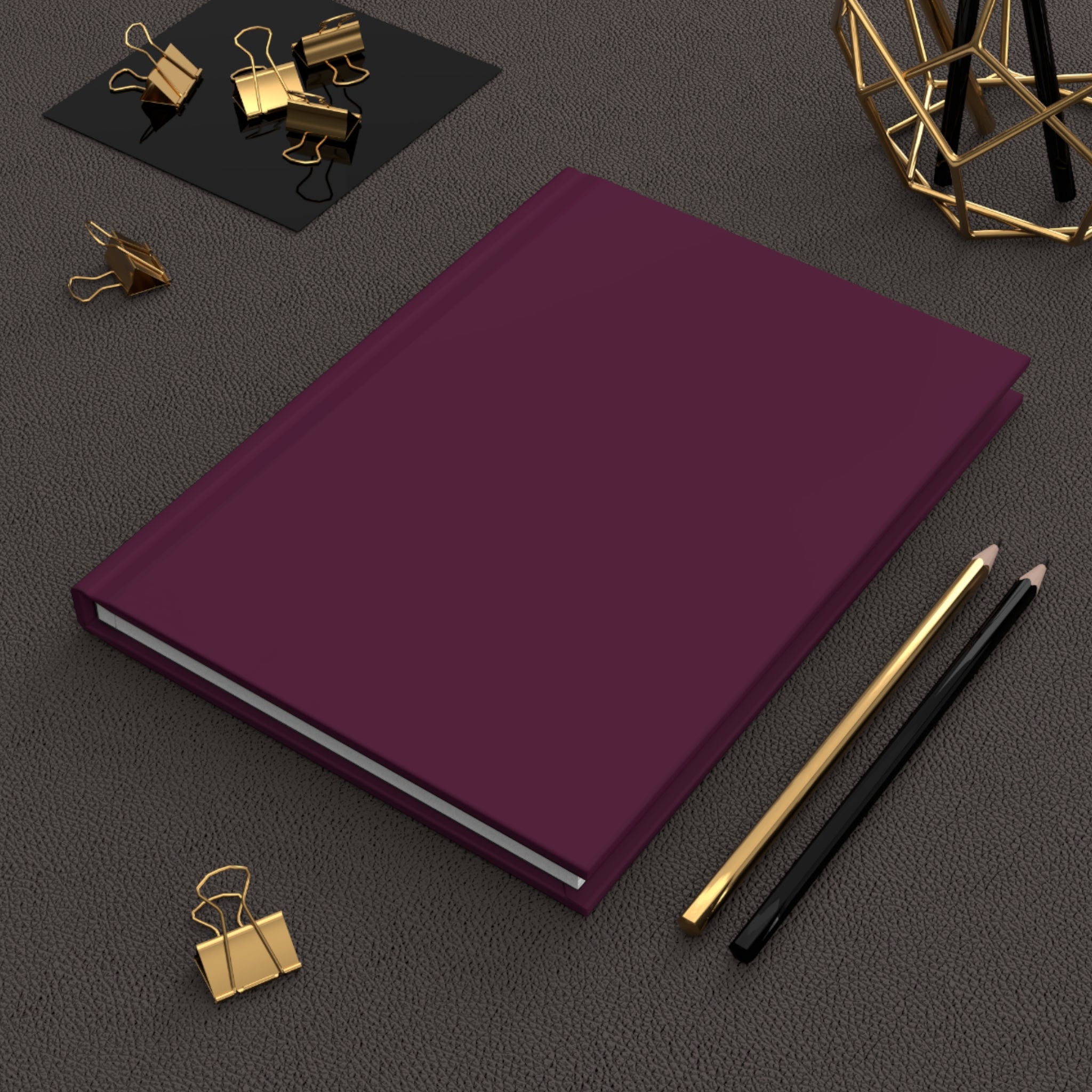 A hardcover notebook or journal in deep plum purple, styled on a textured dark gray surface with gold and black office accessories.