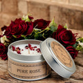 Candle in a metal tin with rose petals surrounded by red roses on a wooden surface.
