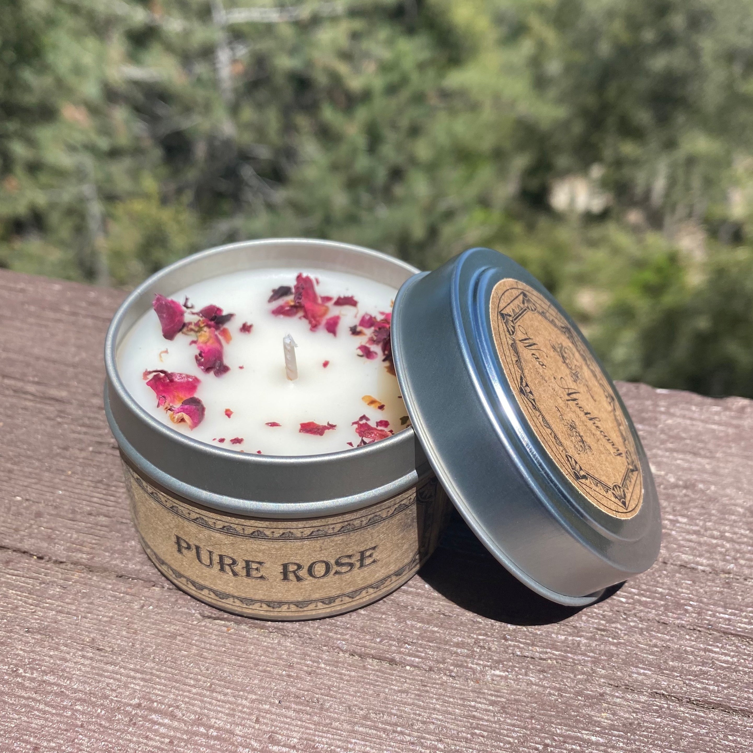 Candle in a tin labeled 'Pure Rose' with rose petals on a wooden surface outdoors.