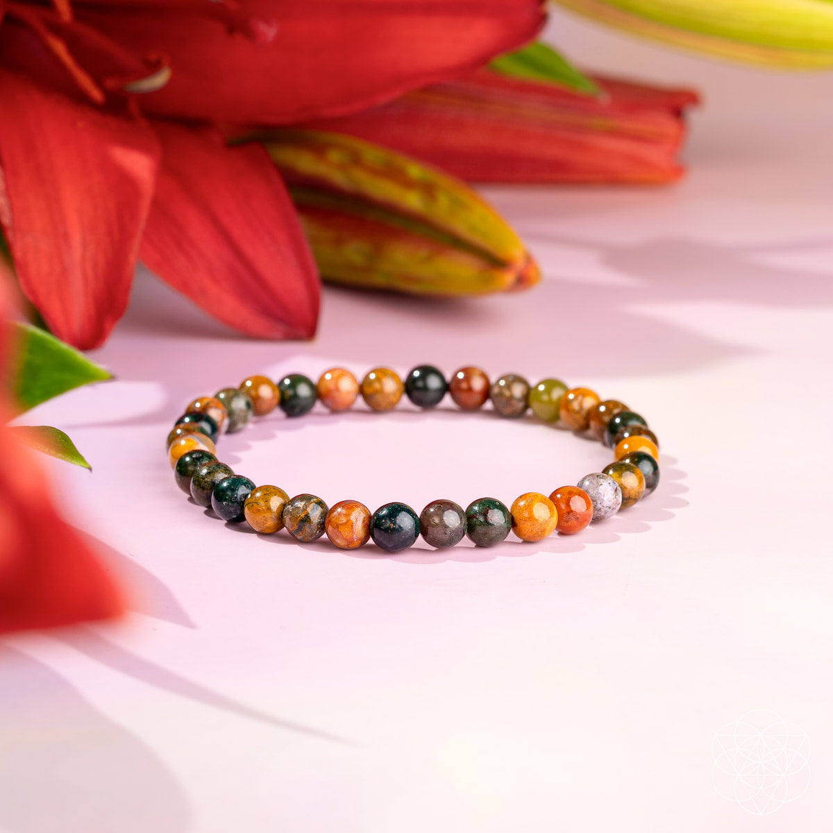 A beaded stretch bracelet made of polished round gemstone beads in earthy orange, brown, green, gray, and black, displayed on a light pink surface with blurred red flowers in the background.