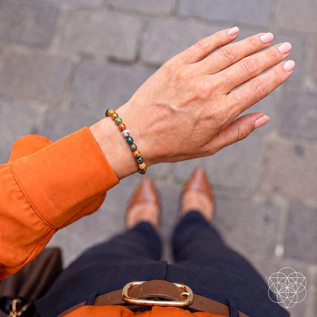 A close-up lifestyle shot of a person's hand wearing a beaded bracelet, with their orange sleeve, navy pants, and brown shoes visible against a gray stone pavement background.
