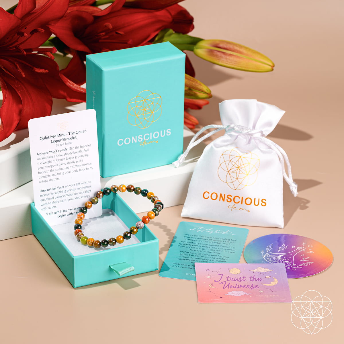 A Conscious brand self-care gift set arranged on a beige surface with red flowers in the background. The set includes a beaded bracelet in an open turquoise box, a turquoise branded box, a white drawstring pouch, a printed card, a circular iridescent sticker, and a pink affirmation card.