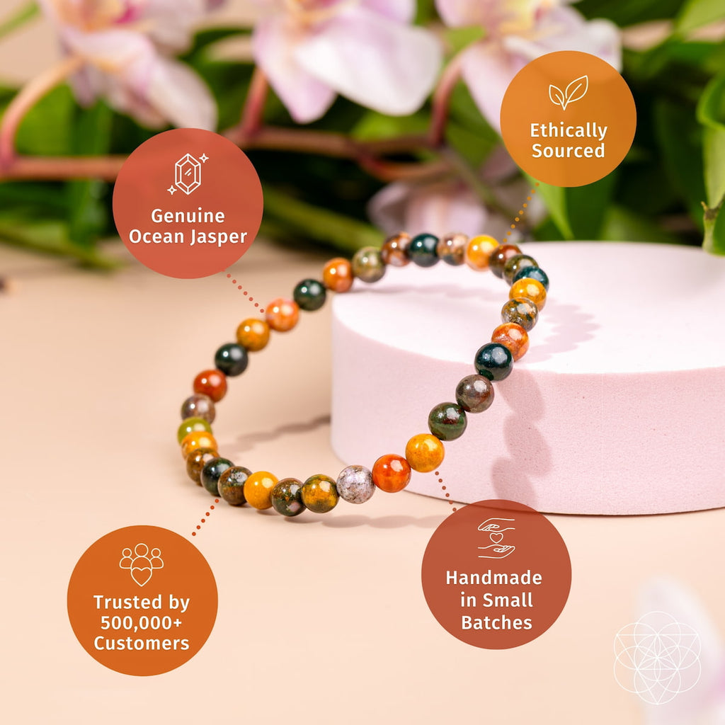 A beaded stretch bracelet made of polished ocean Jasper stones in orange, green, brown, and dark gray, displayed on a light pink cylindrical pedestal. The background is a soft beige with blurred pink flowers and green leaves.