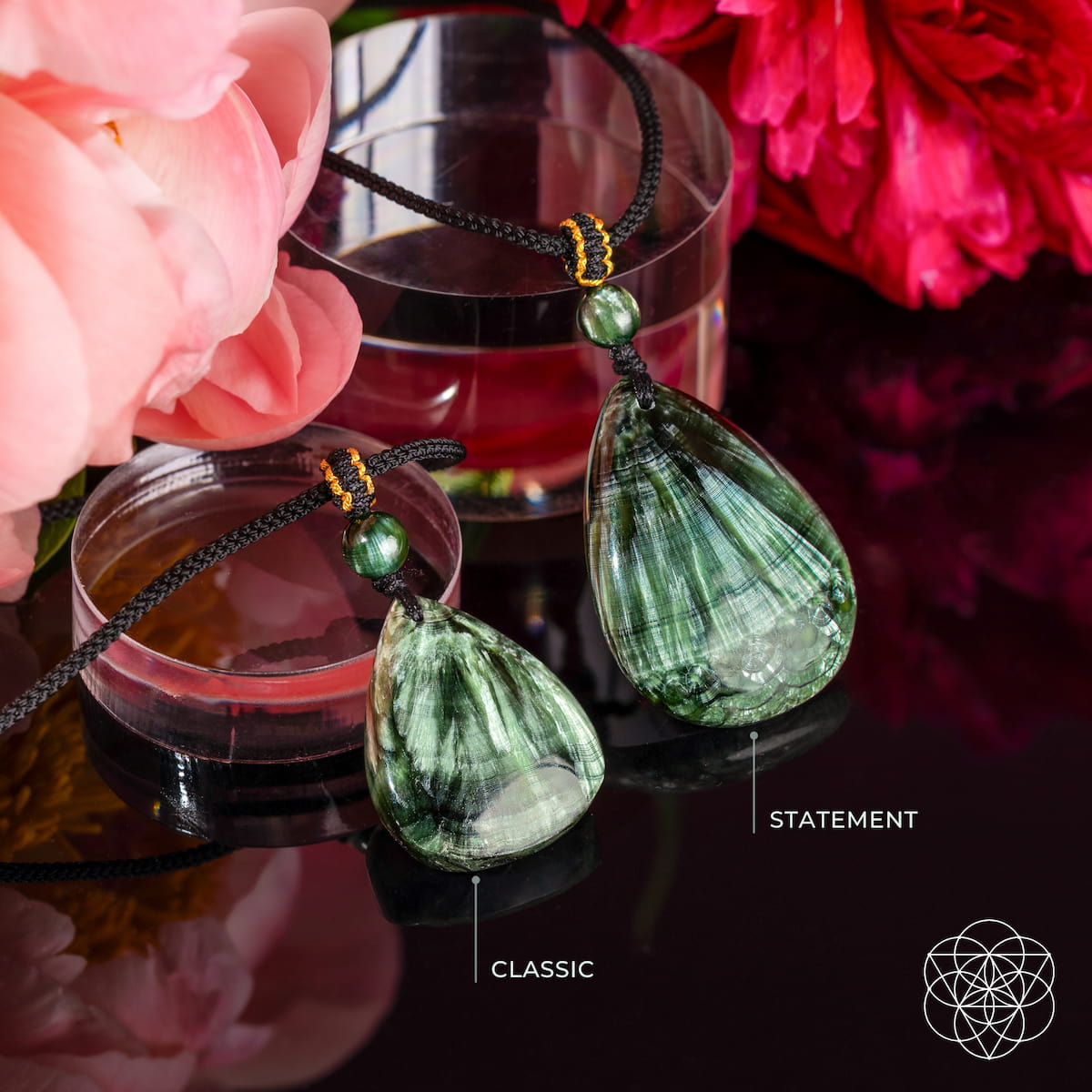 Two green teardrop pendant necklaces with black cords and small green beads, displayed on a reflective dark surface with pink flowers and a glass in the background. One pendant is labeled 'CLASSIC' and the other 'STATEMENT'.