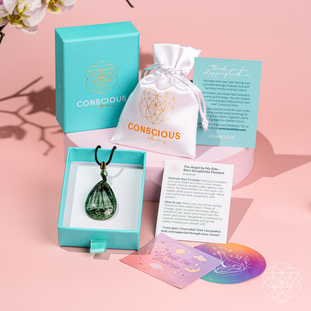 A Conscious Energy jewelry set displayed on a pink background, featuring a green teardrop crystal pendant in a light blue box, a white drawstring pouch, a turquoise gift box, and printed cards and a rainbow coaster.
