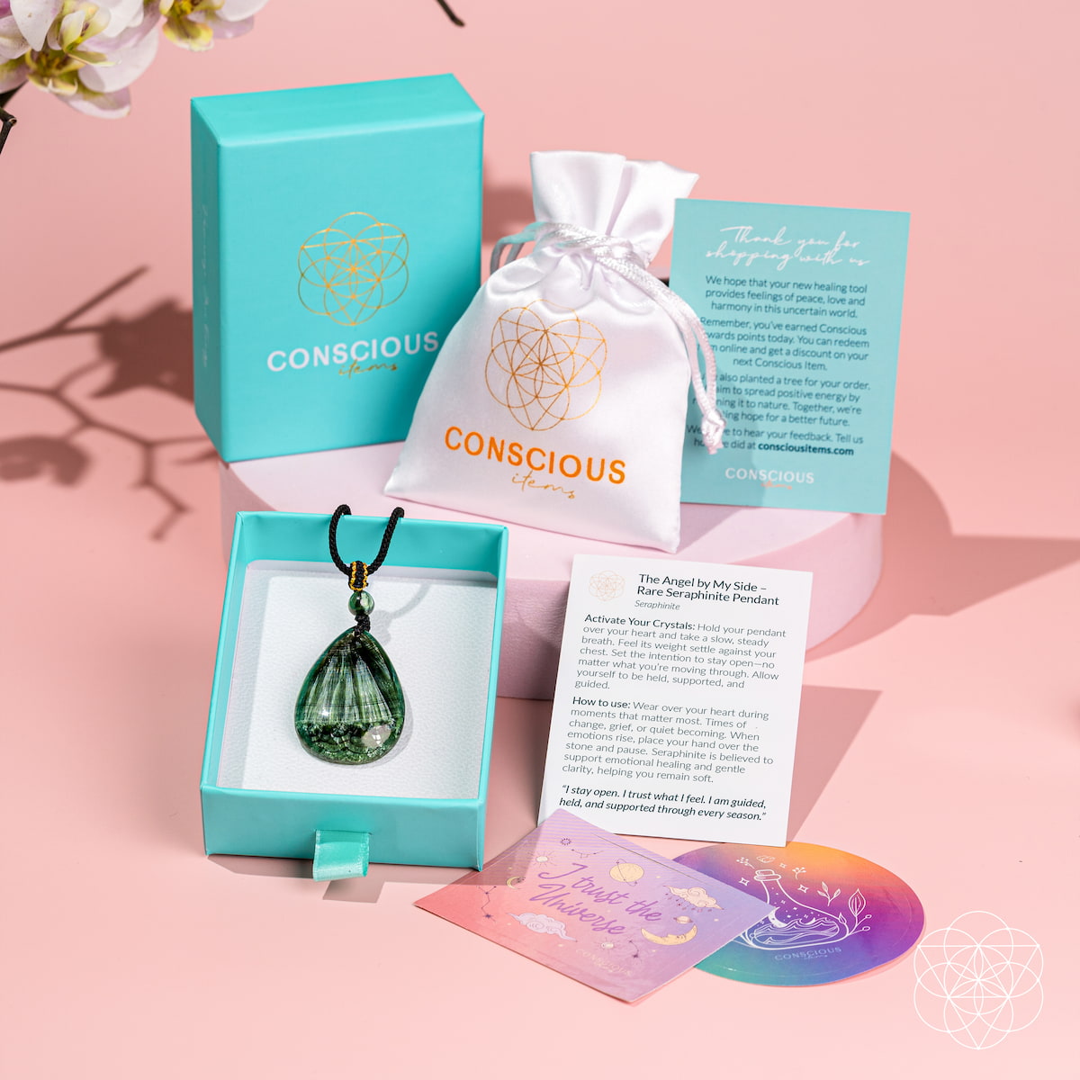 A Conscious Energy jewelry set displayed on a pink background, featuring a green teardrop crystal pendant in a light blue box, a white drawstring pouch, a turquoise gift box, and printed cards and a rainbow coaster.
