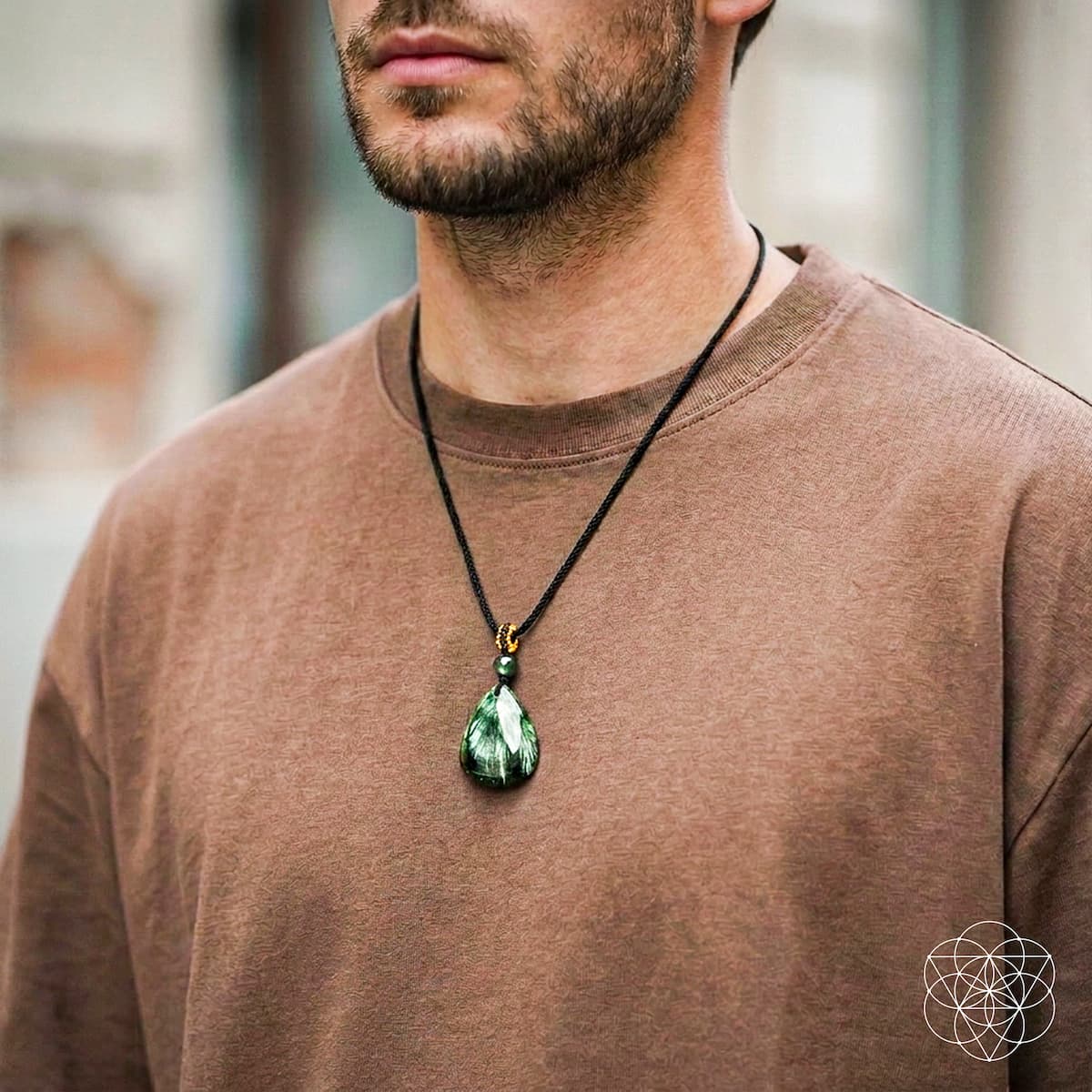 A man wearing a brown crewneck sweatshirt and a black cord necklace with a green teardrop pendant featuring gold metal accents.