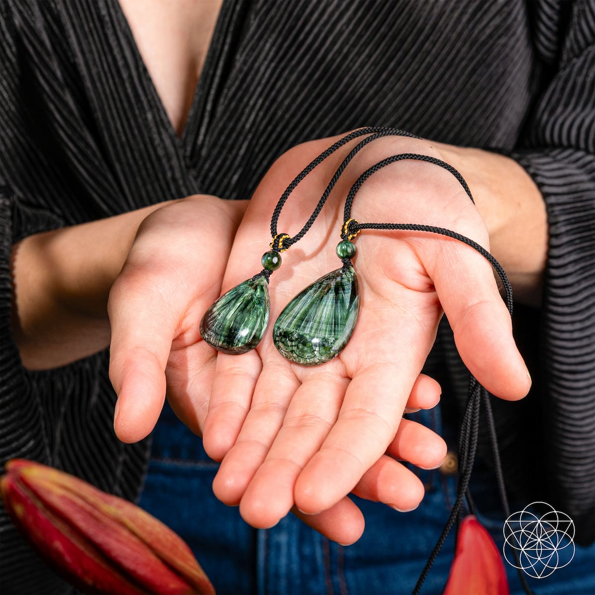 Two green teardrop-shaped crystal pendants on black cords, held in a person's hands. The person is wearing a black ribbed cardigan and blue jeans, with red flowers in the foreground.