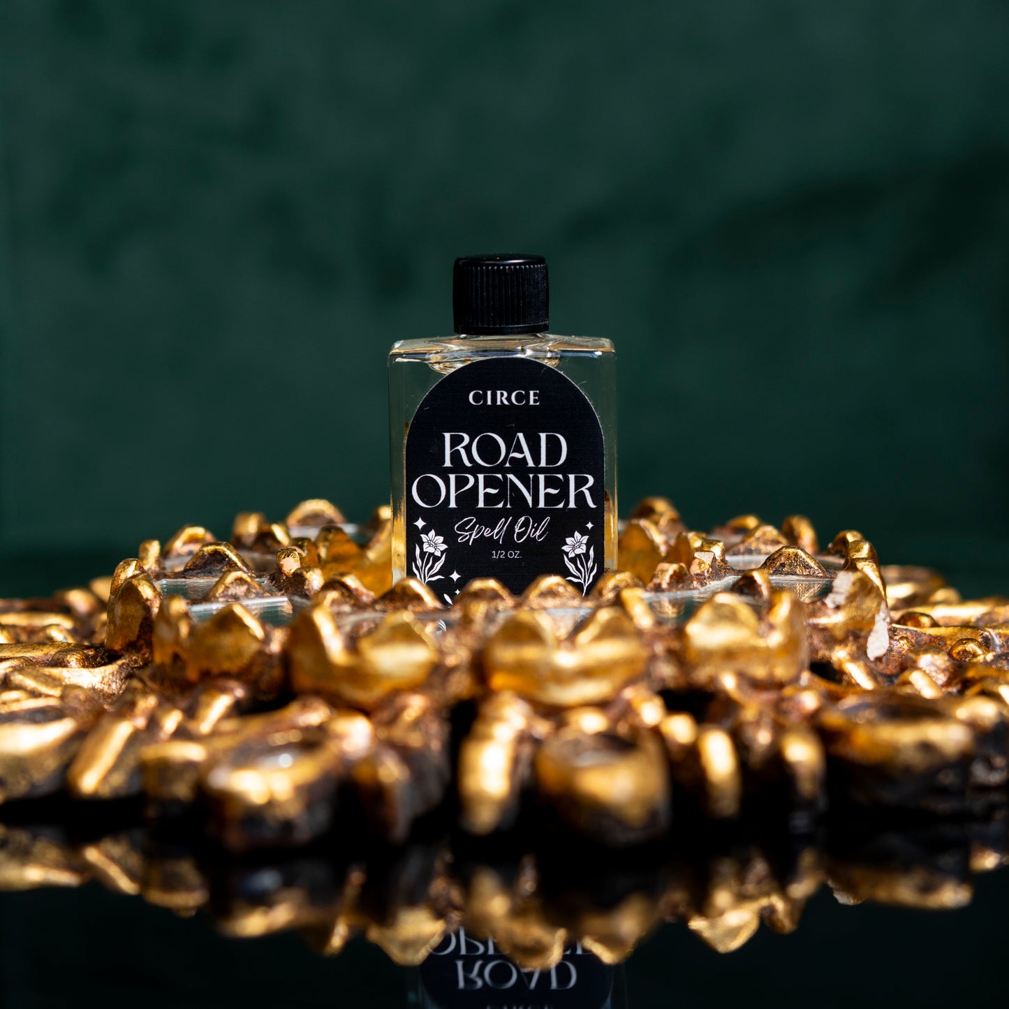 Bottle of Circe Road Opener Spell Oil surrounded by gold coins on a reflective surface with a dark background.