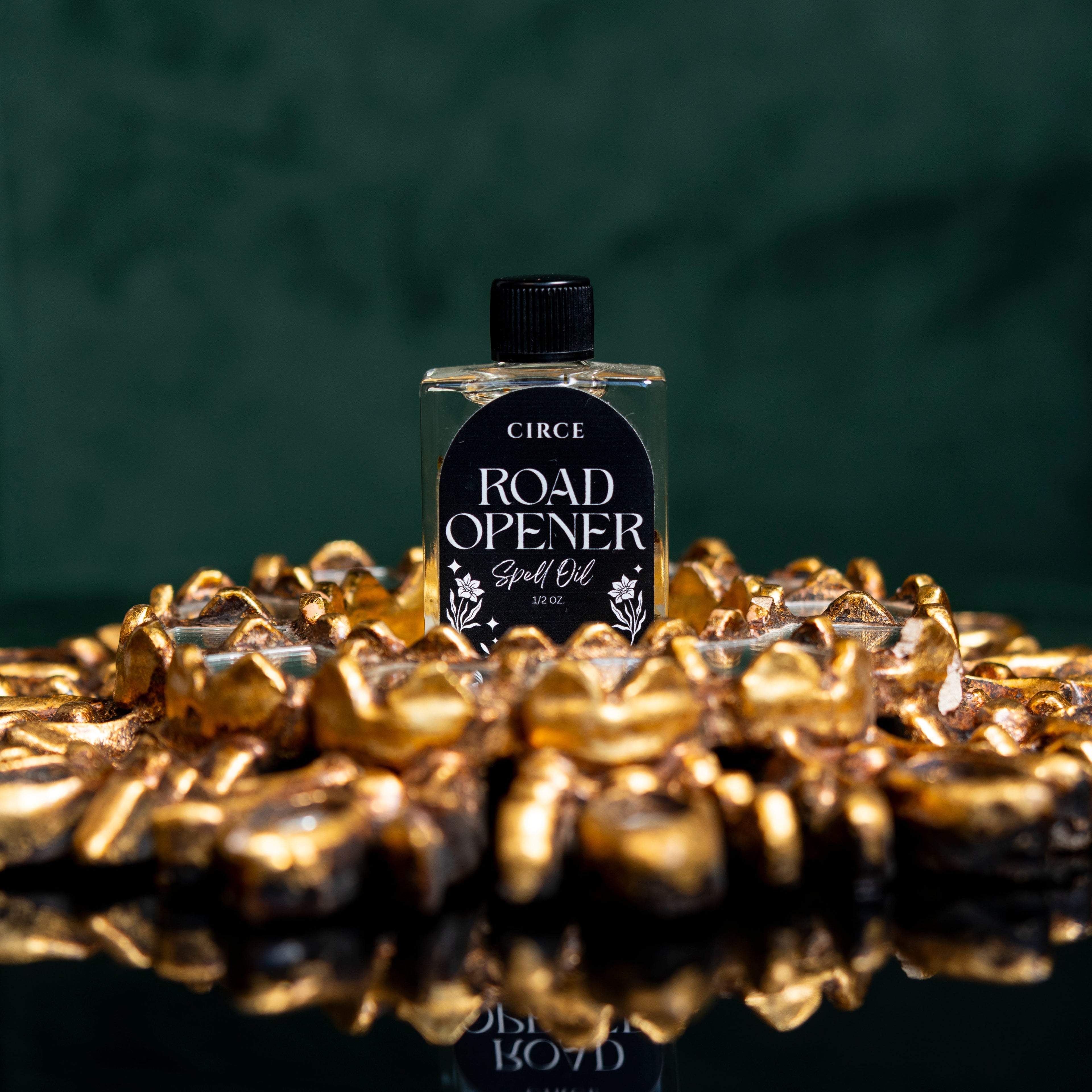 Bottle of Circe Road Opener Spell Oil surrounded by gold coins on a reflective surface with a dark background.