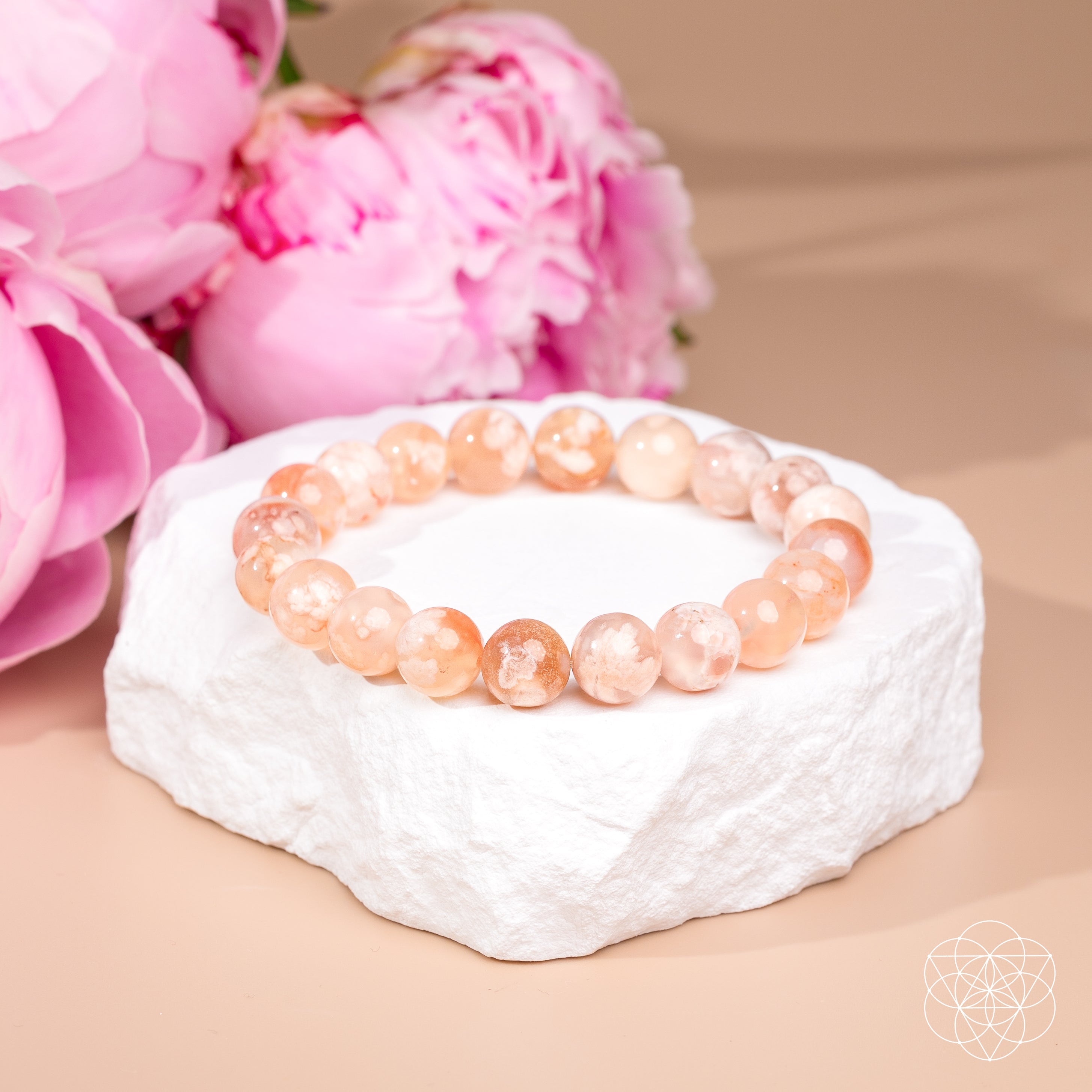 A beaded stretch bracelet made of peachy pink translucent gemstone beads, displayed on a white textured stone riser with pink flowers in the background.