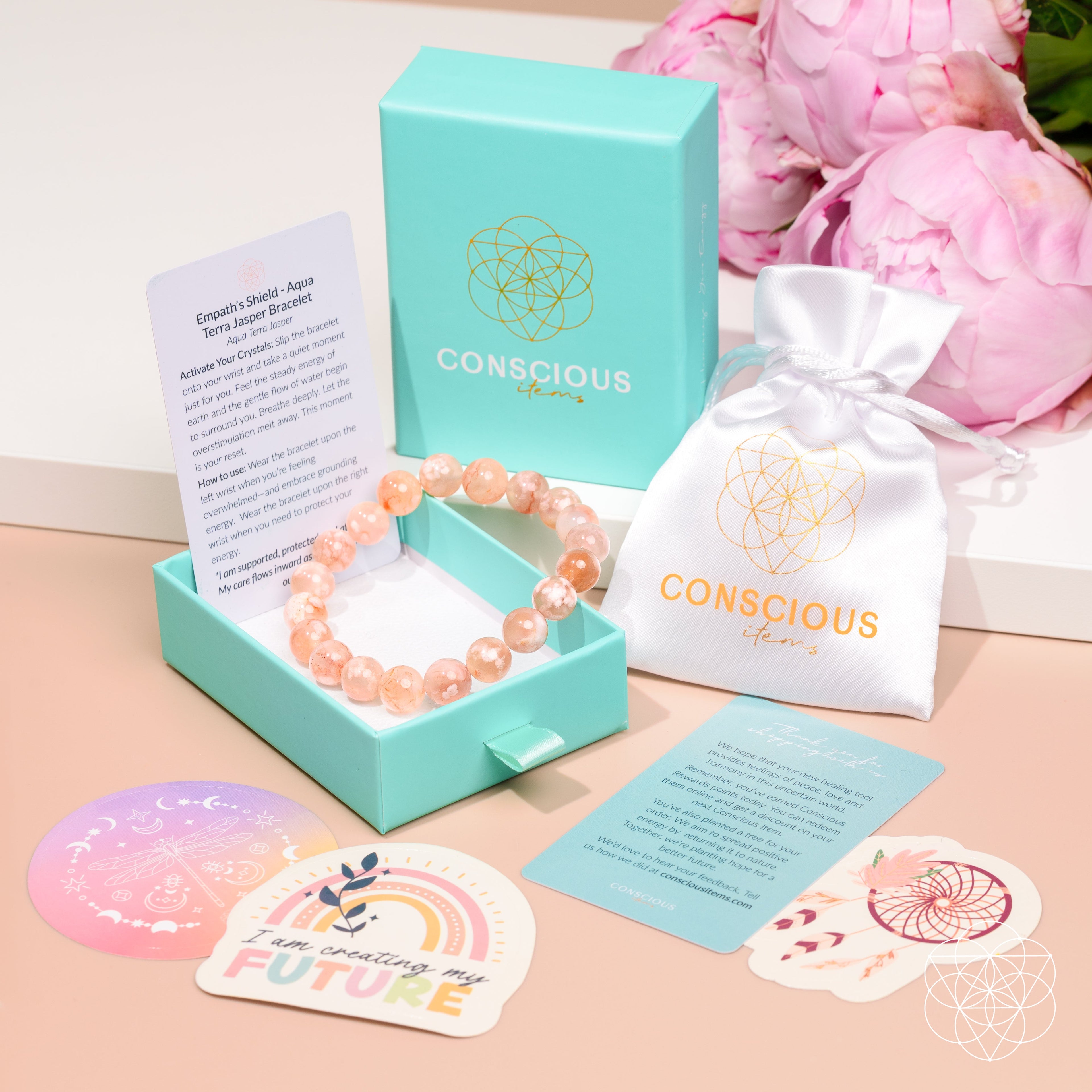 A Conscious Energy brand gemstone bracelet in a peachy-orange hue, displayed inside its mint green presentation box next to a white drawstring pouch, cards, and a pink flower arrangement in the background.