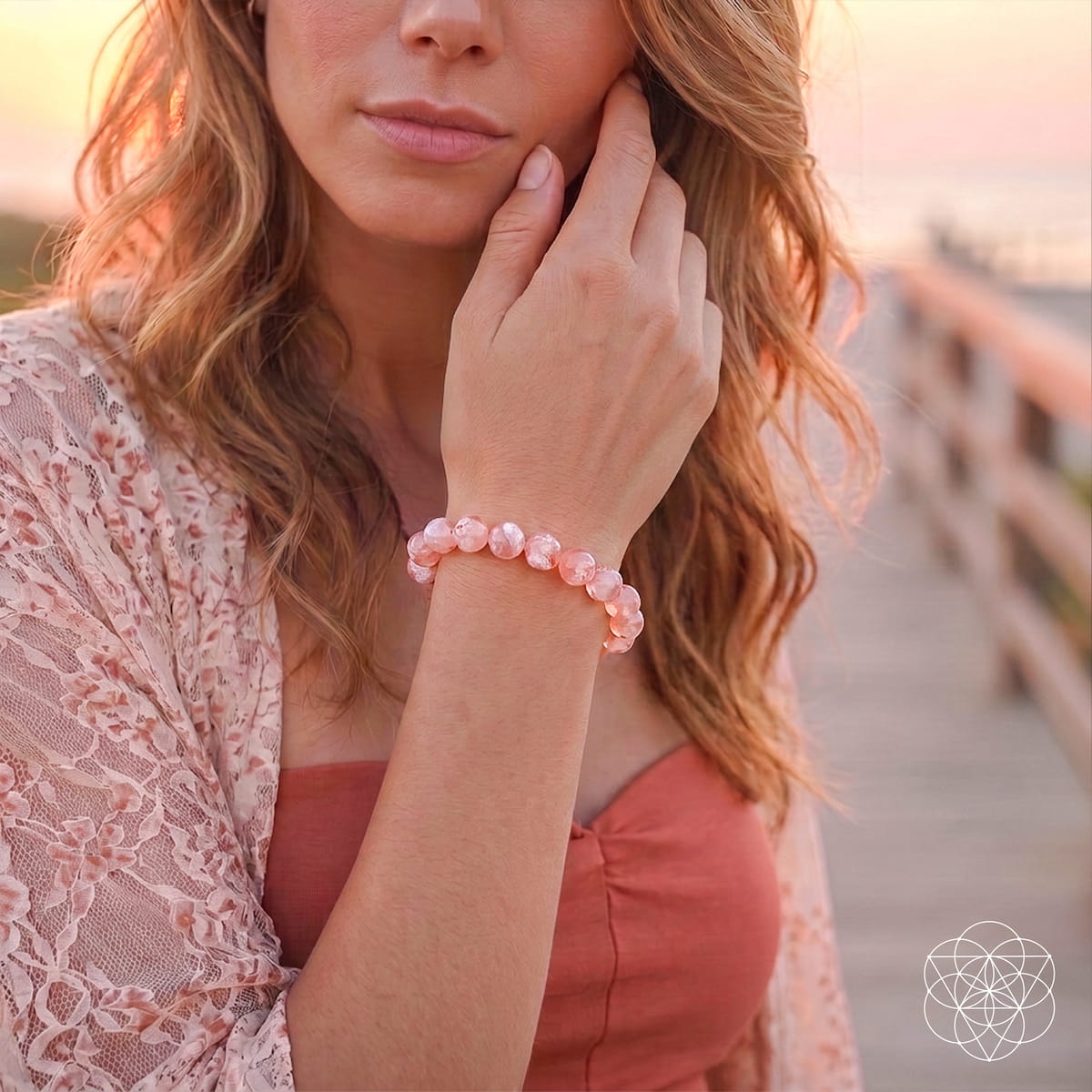 A woman outdoors at sunset wearing a pink beaded bracelet on her wrist, with a lace cardigan and coral top visible.