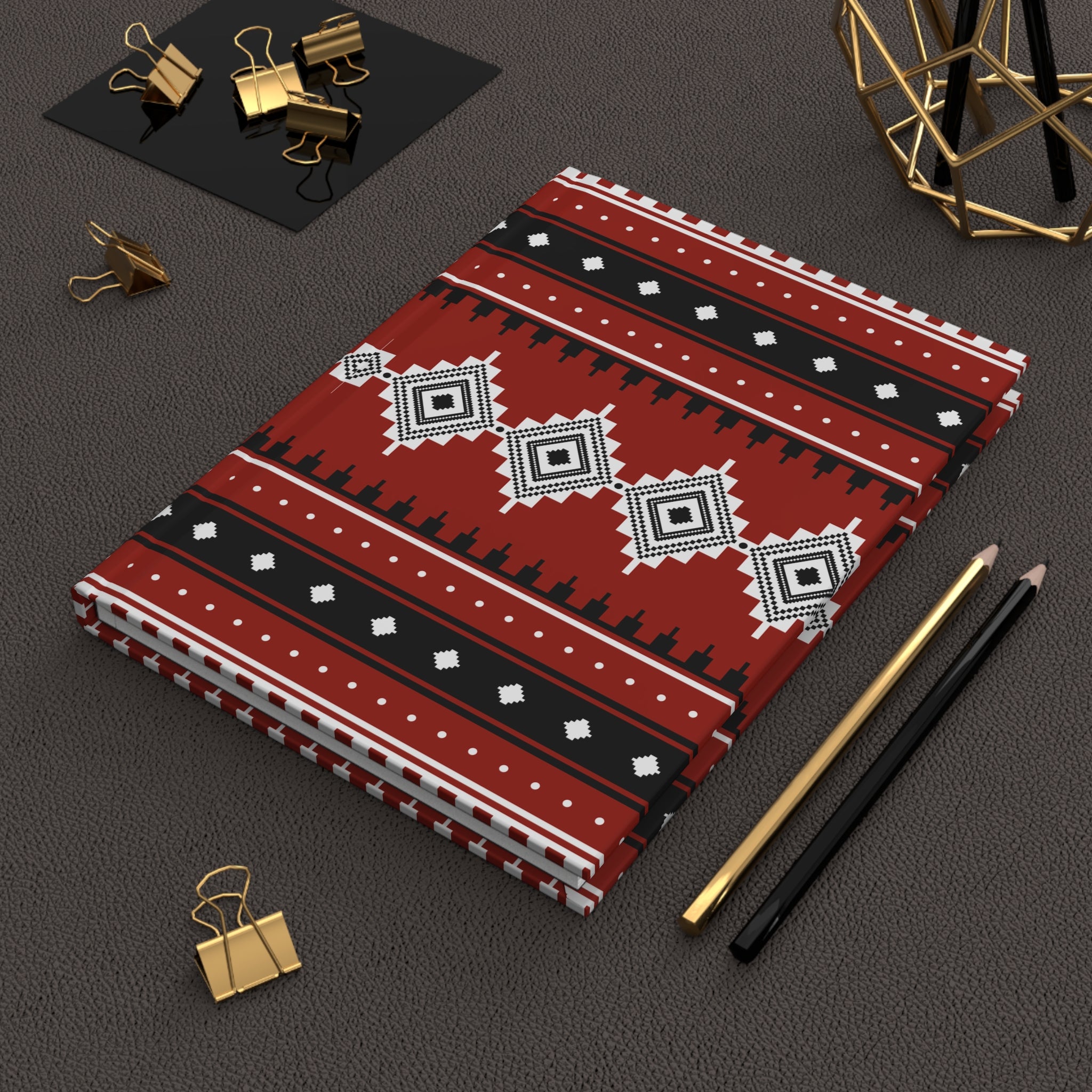 A stack of patterned notebooks or journal covers with a red, black, and white geometric tribal design, styled on a dark textured surface with gold binder clips, pencils, and a gold geometric decor object.