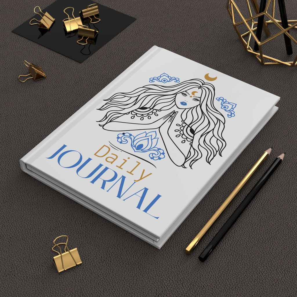 A white hardcover daily journal with a black line-art illustration of a woman with long flowing hair, blue decorative motifs, and the words 'Daily JOURNAL' in blue and gold, styled on a dark textured surface with gold binder clips, pencils, and a geometric decor object.
