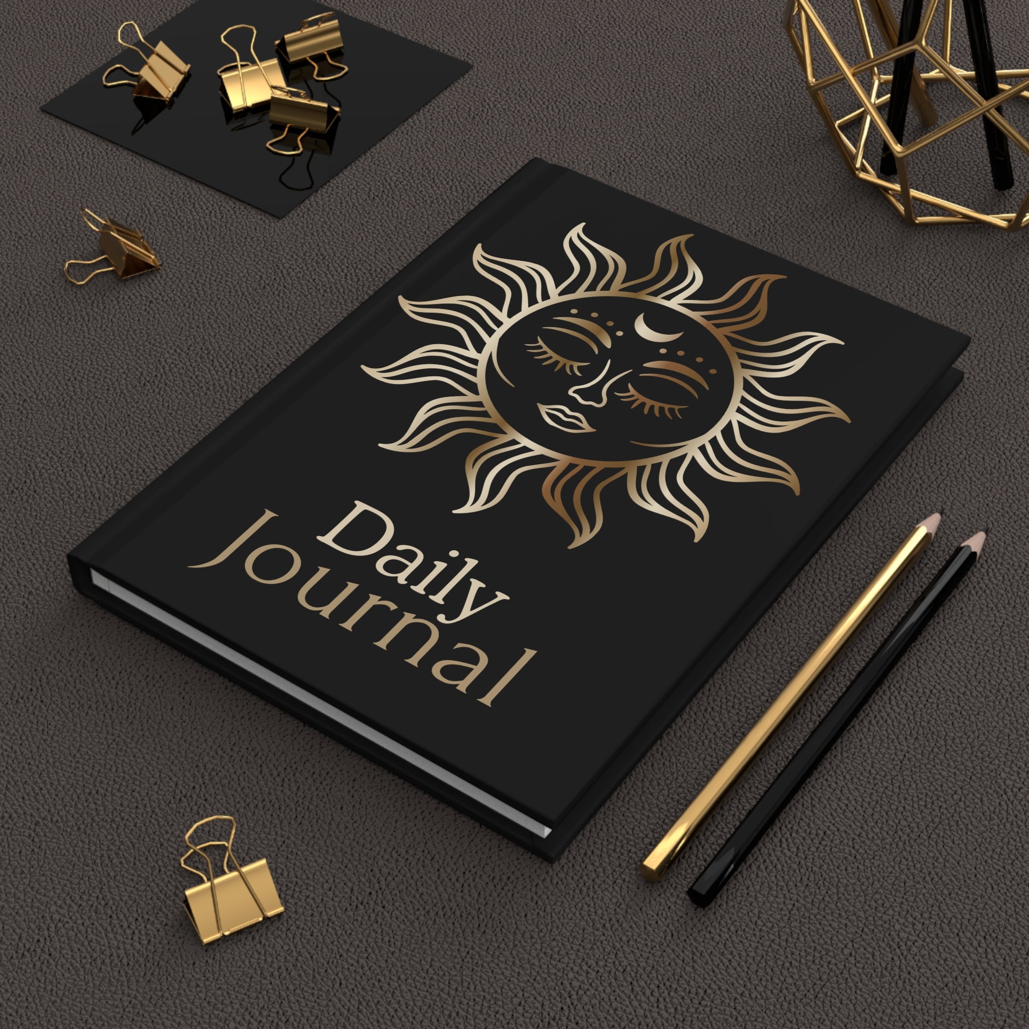 A black hardcover daily journal with a gold sun face illustration and the words 'Daily Journal' on the cover, styled on a dark textured surface with gold and black pencils, binder clips, and a geometric decor object.