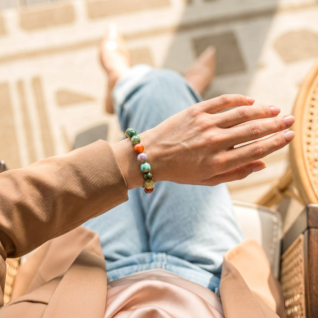 A person wearing a beaded bracelet with green, orange, and white beads, sitting in a sunlit outdoor chair with their legs crossed.