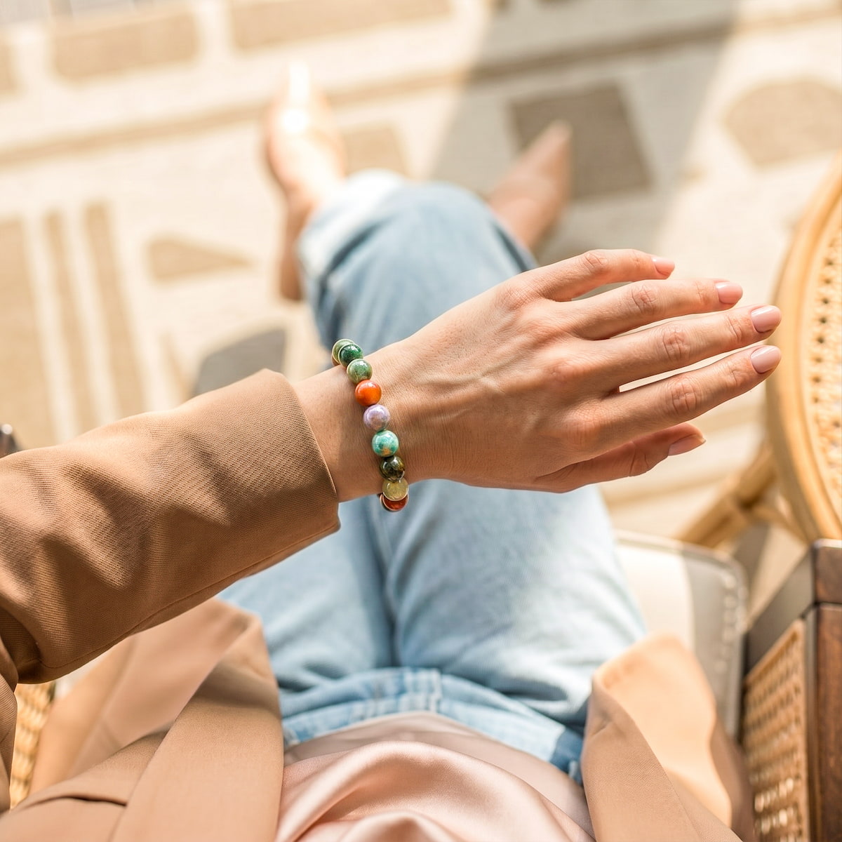 A person wearing a beaded bracelet with green, orange, and white beads, sitting in a sunlit outdoor chair with their legs crossed.