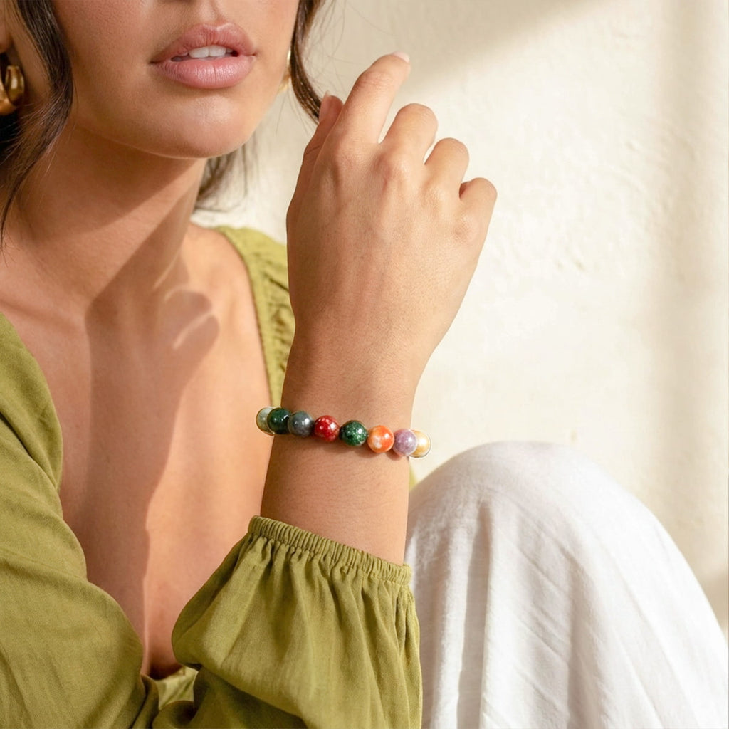 A close-up lifestyle shot of a woman wearing a colorful beaded bracelet on her wrist, wearing an olive green top and white pants against a warm, sunlit cream wall.
