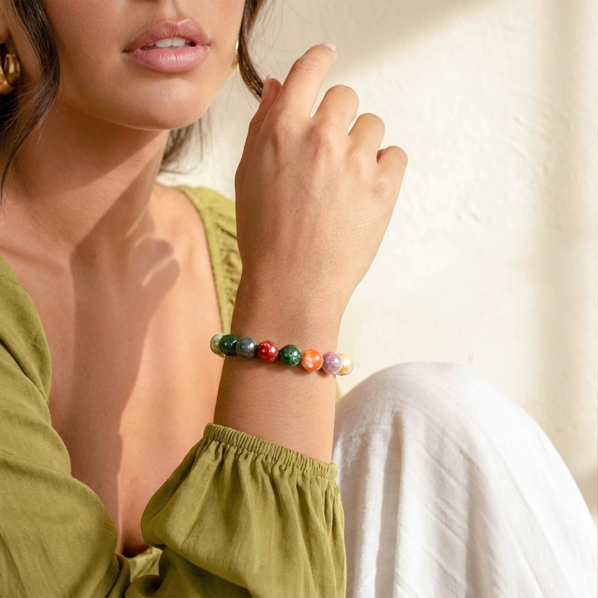A close-up lifestyle shot of a woman wearing a colorful beaded bracelet on her wrist, wearing an olive green top and white pants against a warm, sunlit cream wall.