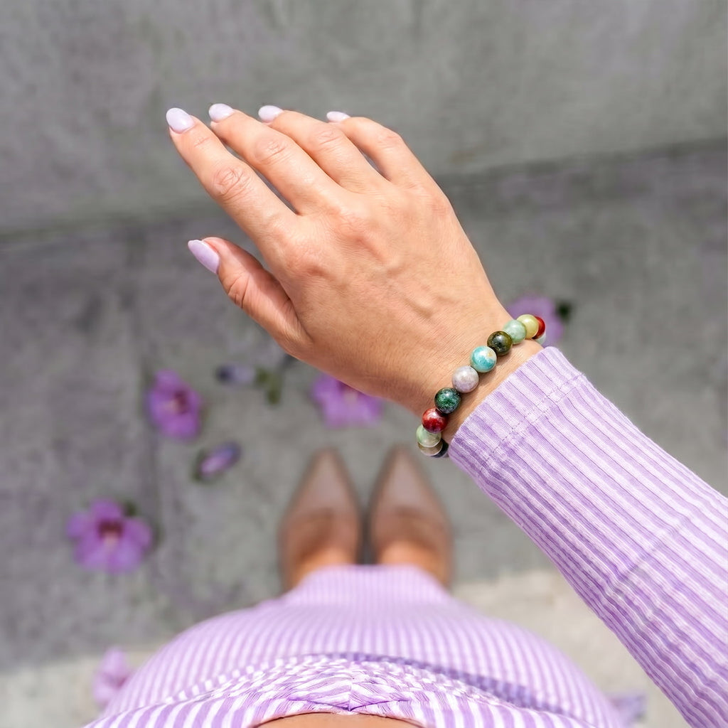 A close-up of a person's hand wearing a beaded bracelet with green, red, white, and light blue beads, styled with a lavender striped long-sleeve top and skirt against a gray concrete background.