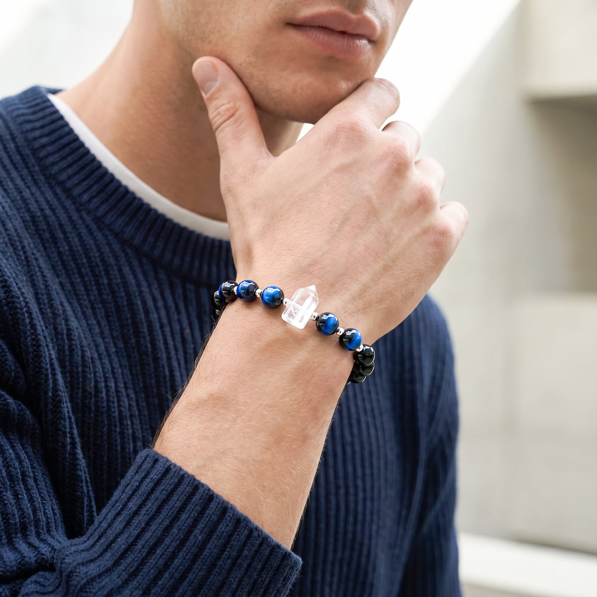 A close-up of a man wearing a dark blue ribbed sweater, showing a beaded bracelet on his wrist. The bracelet features dark blue and black beads with a clear crystal point accent.