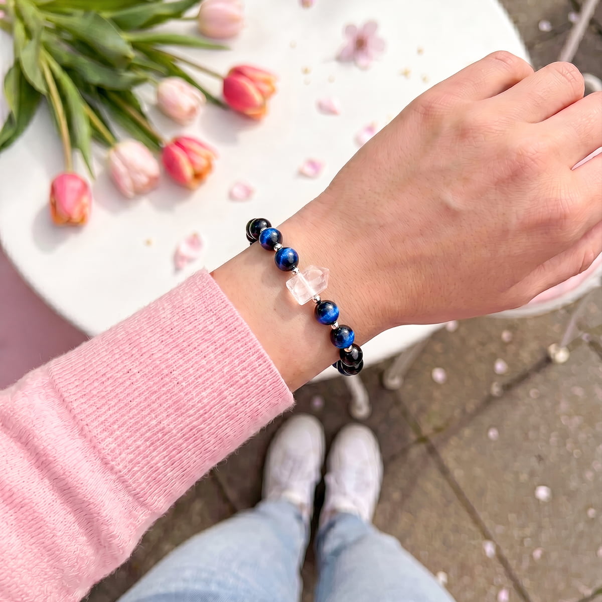 A person wearing a beaded bracelet with blue, black, and clear faceted beads, shown from an overhead perspective on their wrist. They are wearing a pink long-sleeve top, light blue jeans, and white sneakers, with a white table, pink tulips, and petals in the background.