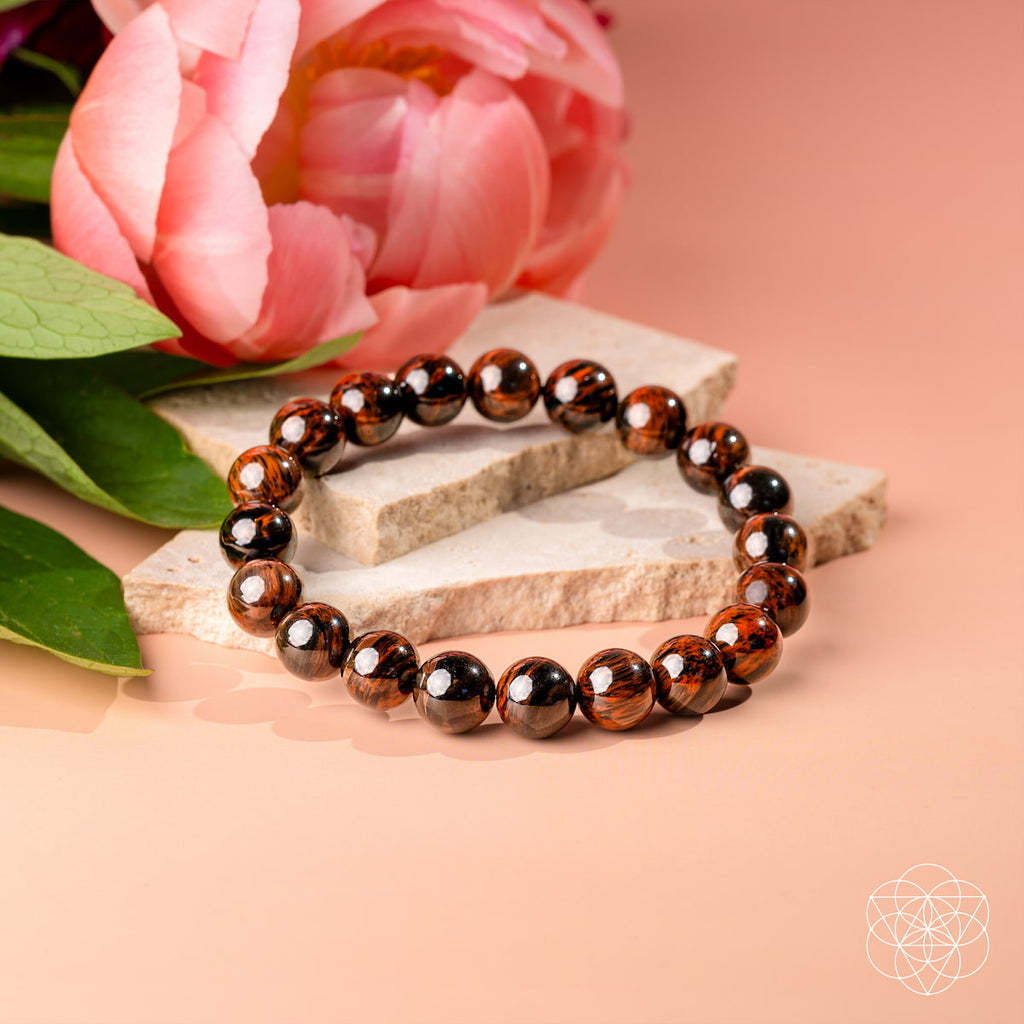A beaded stretch bracelet made of glossy dark brown and black round beads, styled on stacked light stone slabs with a pink flower in the background on a peach-colored surface.