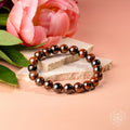 A beaded stretch bracelet made of glossy dark brown and black round beads, styled on stacked light stone slabs with a pink flower in the background on a peach-colored surface.