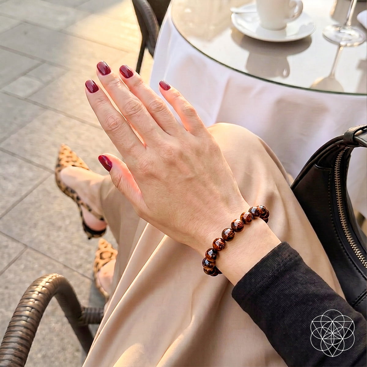 A close-up lifestyle shot of a person's hand and wrist wearing a dark brown beaded bracelet, set against a cafe background with a glass table, white cup, and a black handbag.