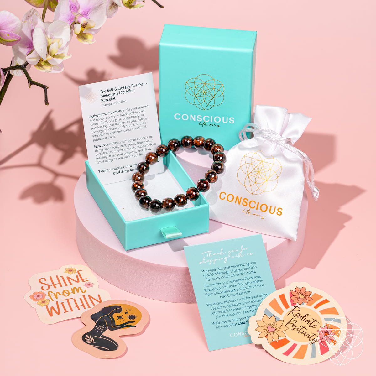 A Conscious Energy self-healing bracelet made of dark brown and amber beads, displayed in a turquoise jewelry box alongside its packaging, including a white drawstring pouch, cards, and stickers on a pink background.