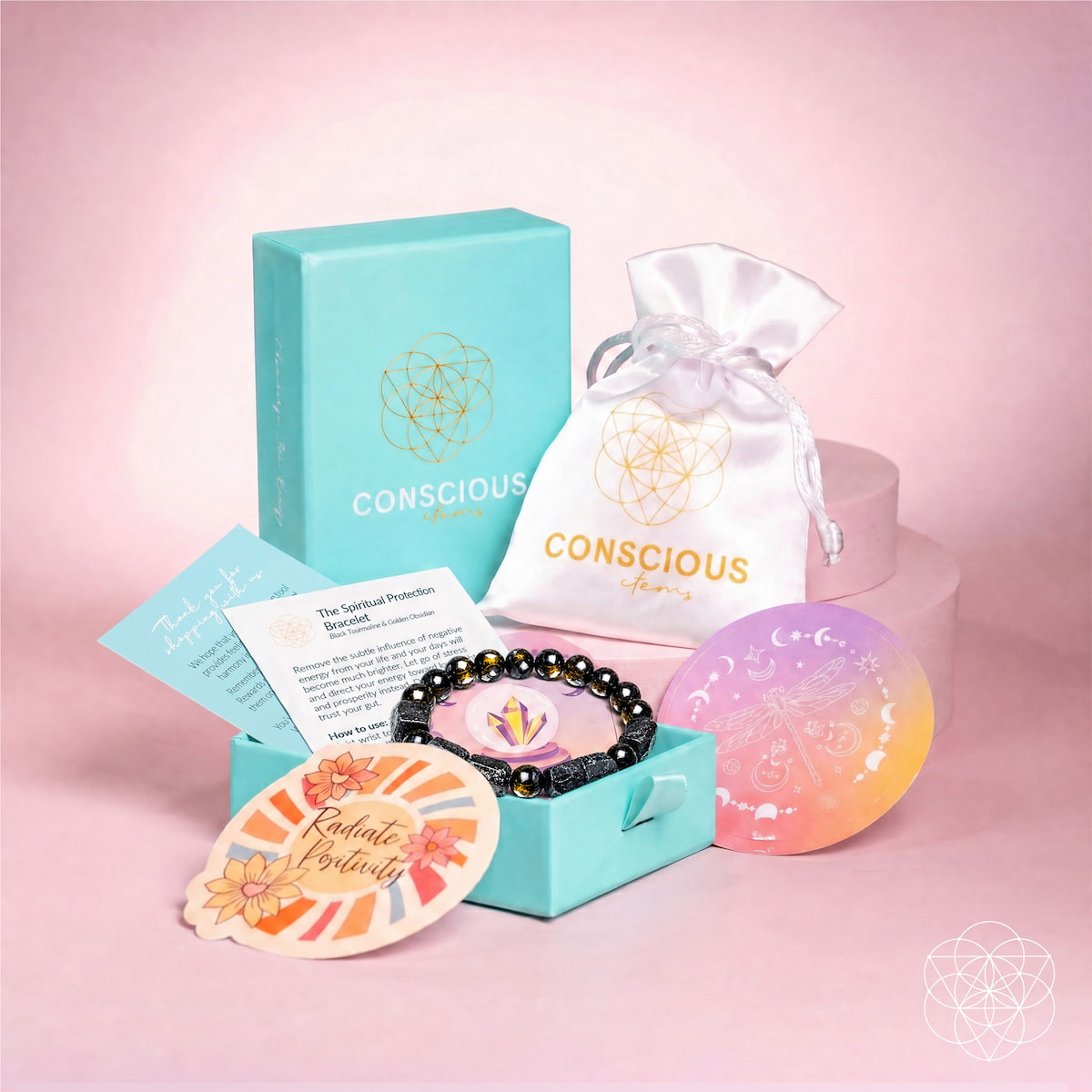 A Conscious Living self-care gift set arranged on a pink studio background. The set includes a turquoise branded box, a white satin drawstring pouch, a black beaded bracelet, a pastel gradient soap bar, a card, and a decorative tag.