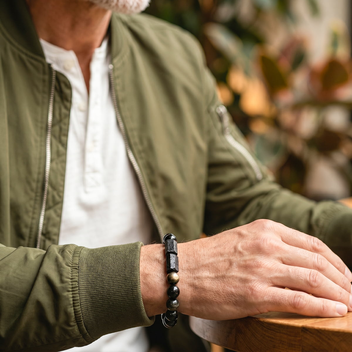 A close-up of a man's hand resting on a wooden table, wearing a black beaded bracelet with polished stone accents. He is wearing an olive green bomber jacket over a white henley shirt, with a softly blurred indoor background.
