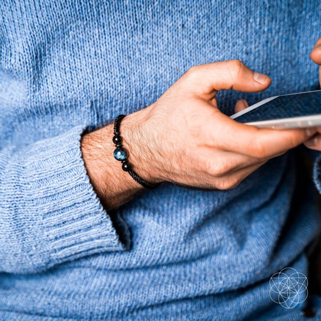 Hand wearing a black beaded bracelet holding a smartphone, with a blue sweater background