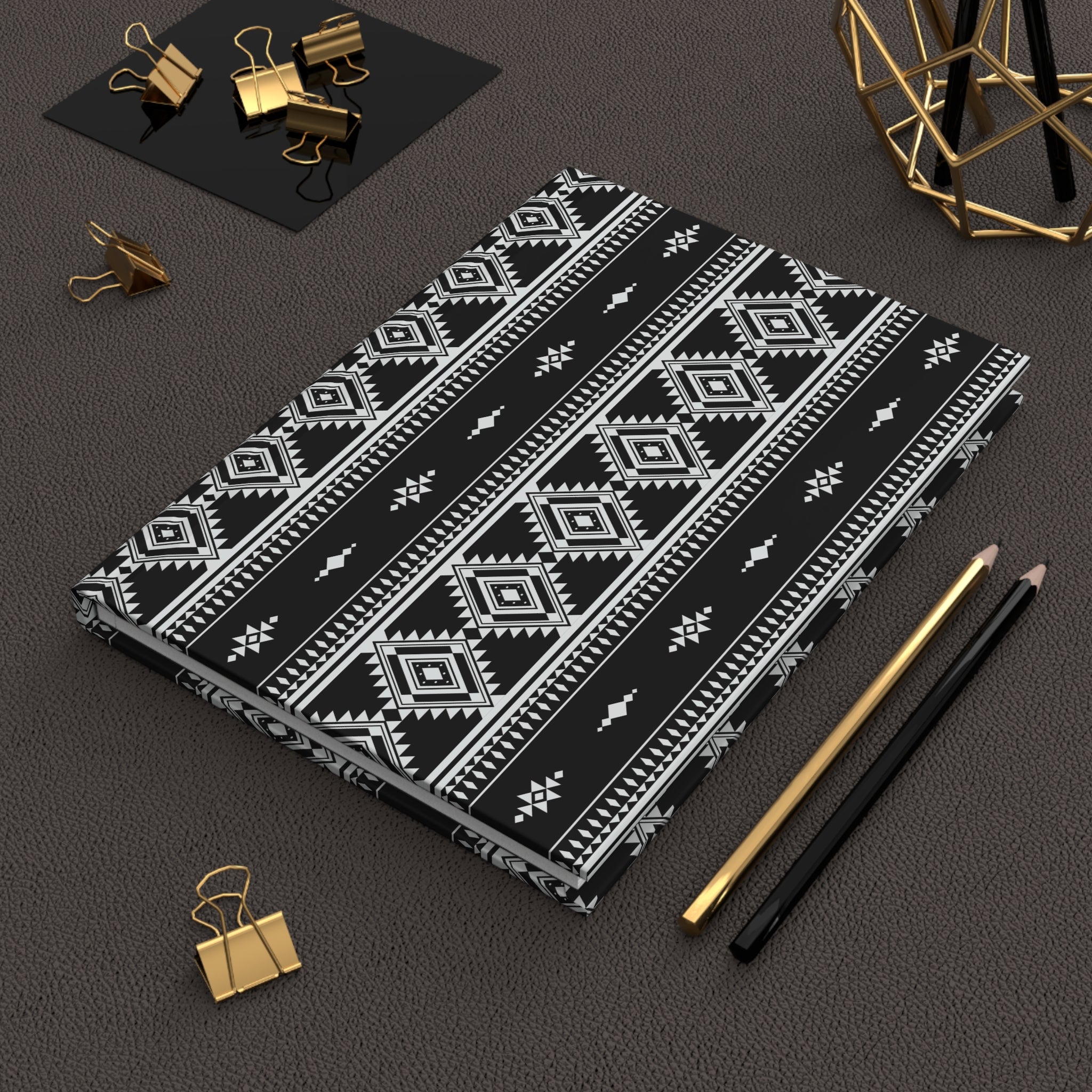 A black notebook or journal with a white geometric tribal pattern, styled on a dark textured surface with gold and black pencils, binder clips, and a gold geometric decor piece.
