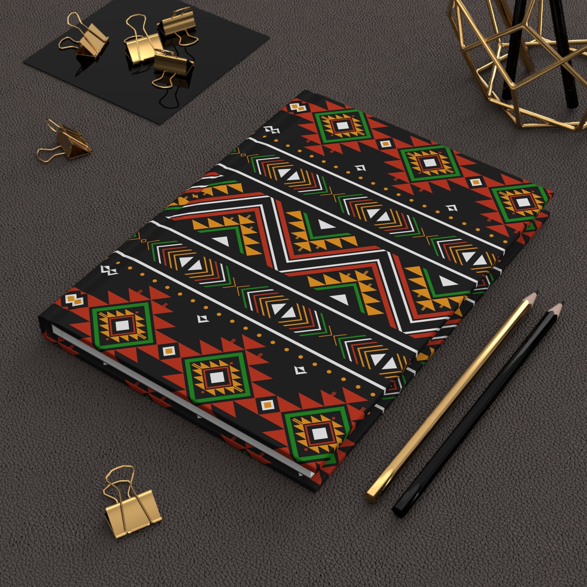 A notebook with a bold geometric tribal pattern in red, green, orange, black, and white, styled on a dark textured surface with gold binder clips, pencils, and a gold geometric decor piece.