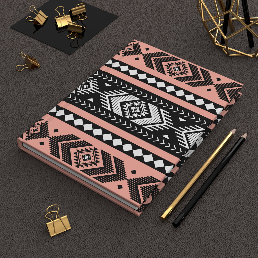 A notebook with a geometric Southwestern-style pattern in pink, black, and white, styled on a dark textured surface with gold and black office accessories.