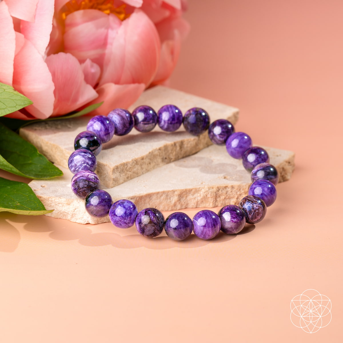 A beaded stretch bracelet made of polished purple gemstone beads with natural inclusions, displayed on stacked stone slabs next to pink flowers on a peach background.