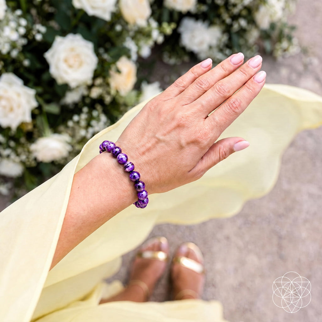 A person's hand and wrist wearing a purple beaded bracelet, with a pale yellow dress, white roses, and gold sandals visible in the background.