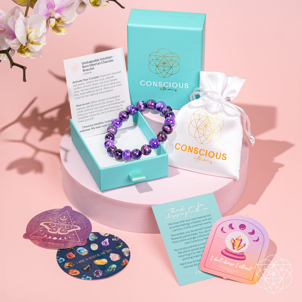 A Conscious Brand crystal charm bracelet in purple beads displayed in a turquoise jewelry box, with its branded turquoise box, white drawstring pouch, instruction card, and two decorative coasters on a pink background.