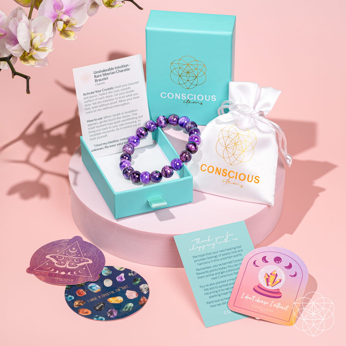 A Conscious Brand crystal charm bracelet in purple beads displayed in a turquoise jewelry box, with its branded turquoise box, white drawstring pouch, instruction card, and two decorative coasters on a pink background.