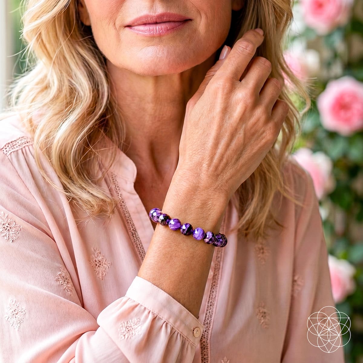 A woman wearing a beaded bracelet made of purple and black marbled beads, paired with a light pink embroidered blouse. The background is a soft-focus garden with pink flowers.