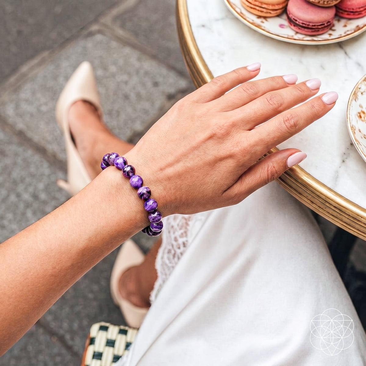 A person wearing a purple beaded bracelet with white pointed-toe heels, seated at a marble table with macarons in the background.