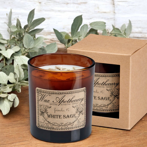 White Sage candle with label and packaging on a wooden surface with greenery