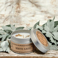 White Sage candle in a metal tin with a wooden label, surrounded by green leaves on a rustic wooden surface.
