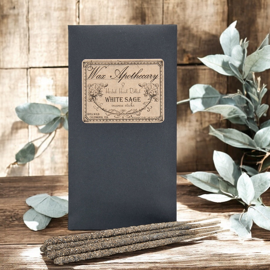 Black box with 'Wax Apothecary' label and white sage incense sticks on a wooden surface with eucalyptus leaves.