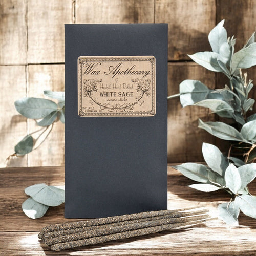 Black box with 'Wax Apothecary' label and white sage incense sticks on a wooden surface with eucalyptus leaves.