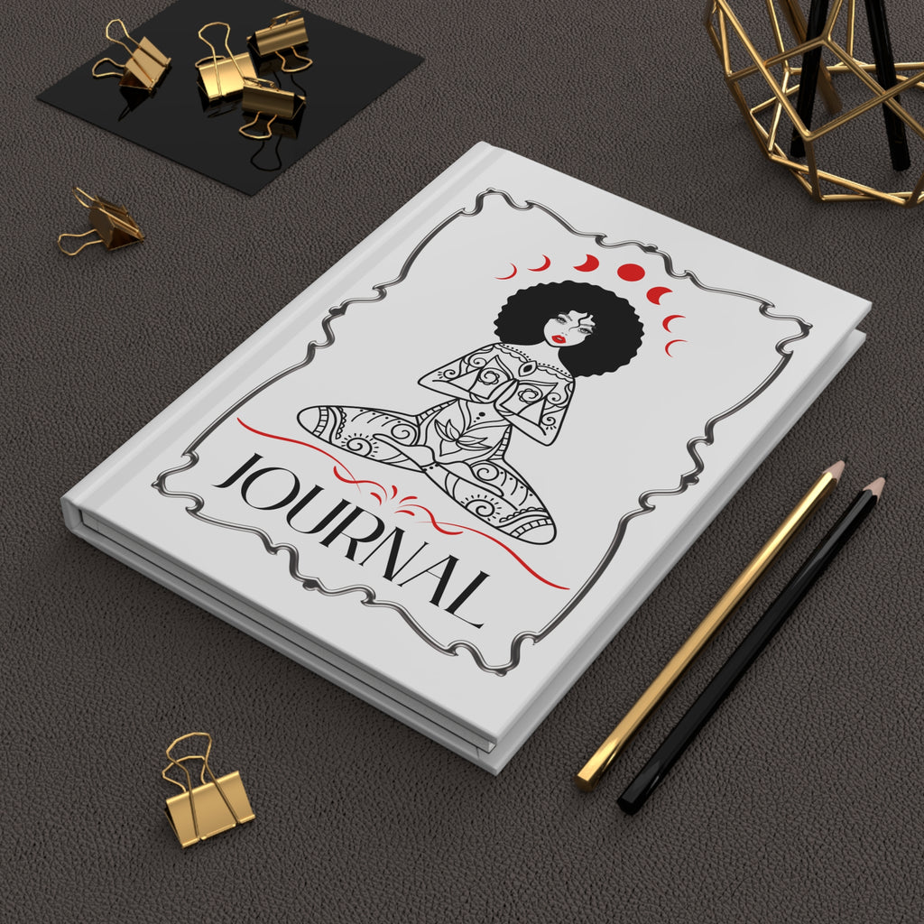 A white hardcover journal with a black and red illustration of a seated woman and the word 'JOURNAL' on the cover, styled on a dark textured surface with gold binder clips, pencils, and a geometric decor object.
